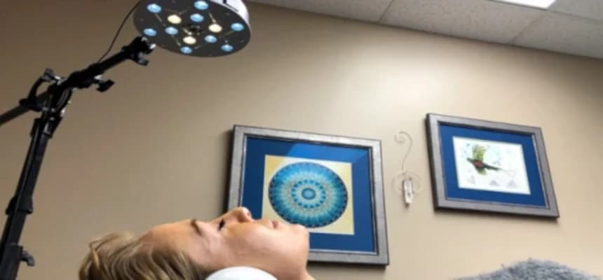 A woman lying, eyes closed, under light therapy equipment emitting a soft glow.